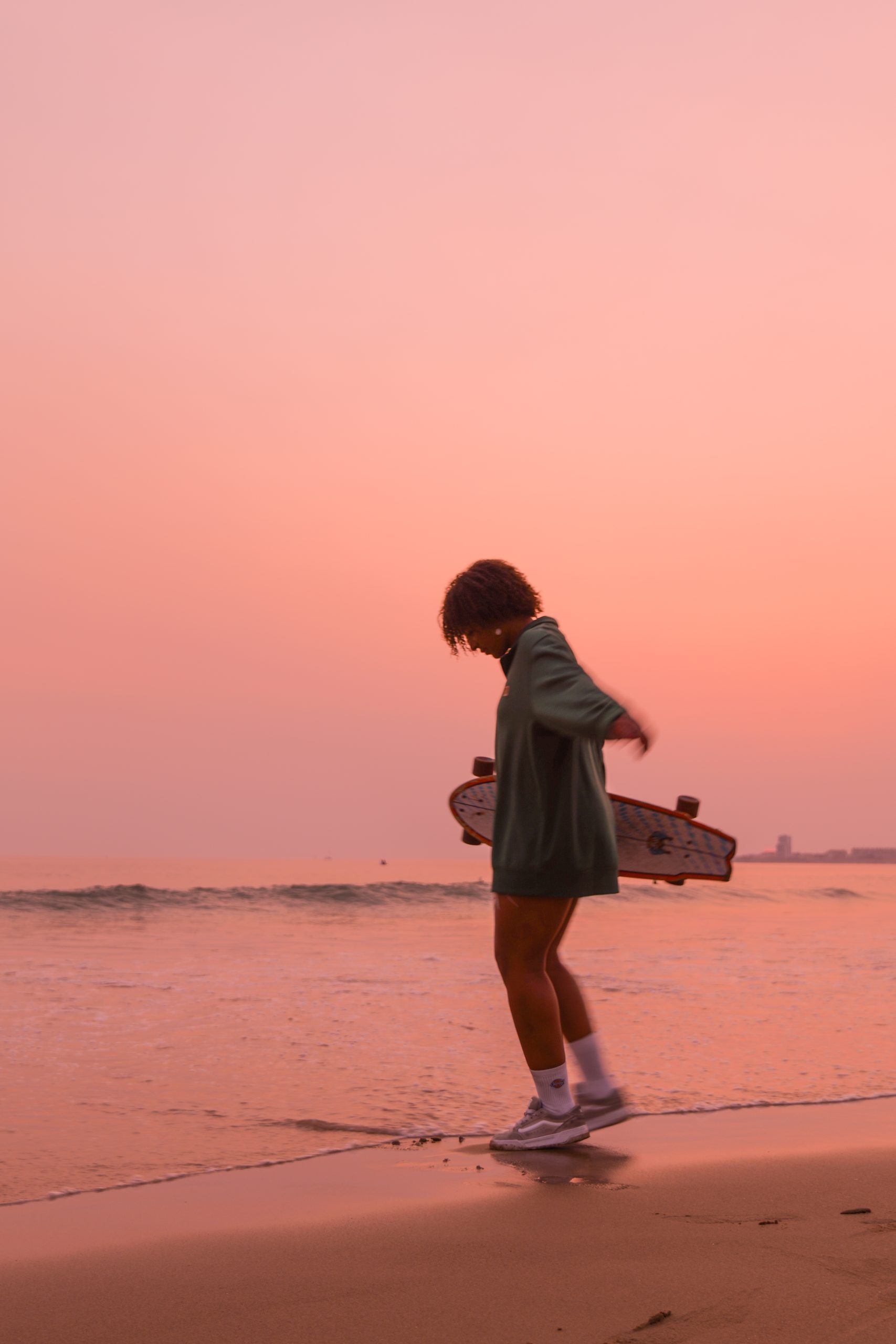 Jeune femme marchant sur le sable au bord de l’océan au coucher du soleil, tenant un skateboard sous le bras, dans une lumière rose et dorée