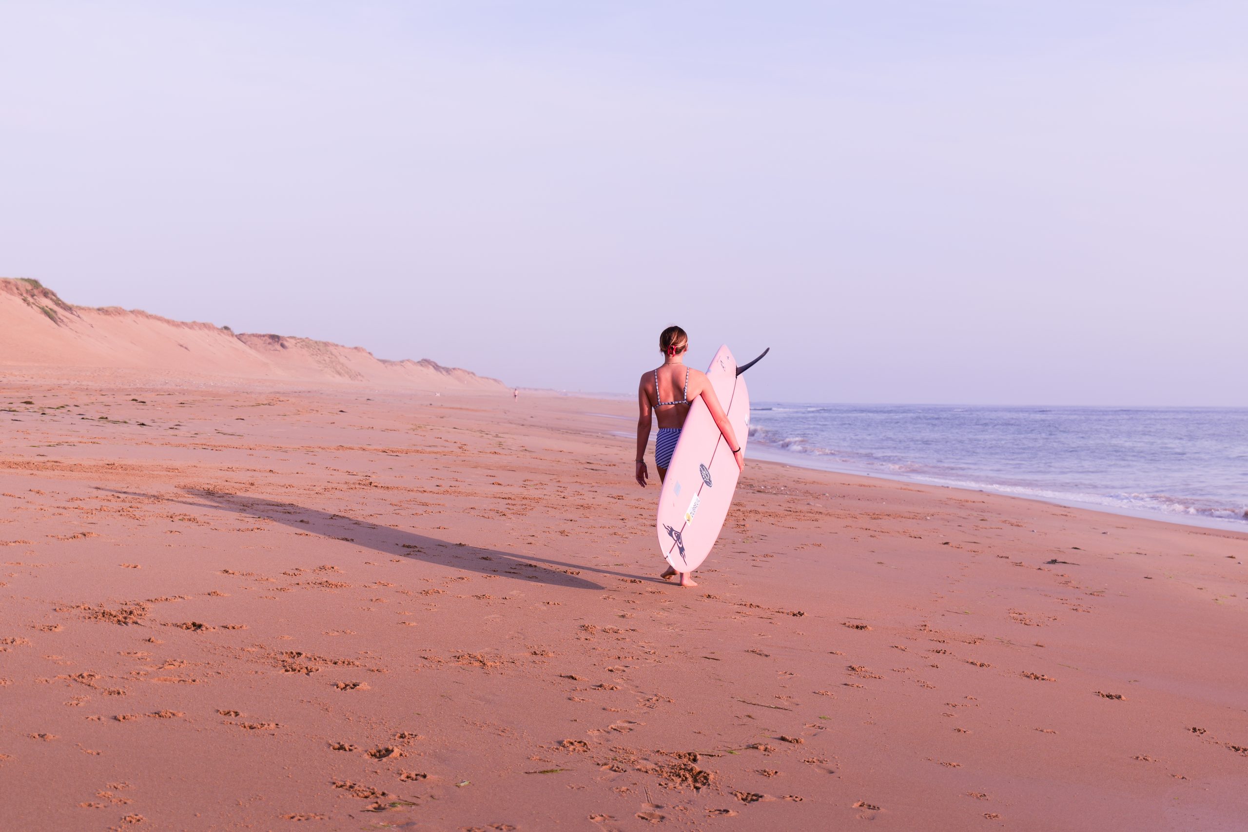 Surfeuse marchant seule sur une plage de sable avec son longboard, face à l’océan, sous une lumière douce de fin de journée.